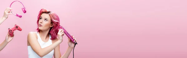 KYIV, UKRAINE - JULY 29, 2020: surprised young woman with colorful hair using straightener near headphones and joystick in hands isolated on pink, panoramic shot — Stock Photo