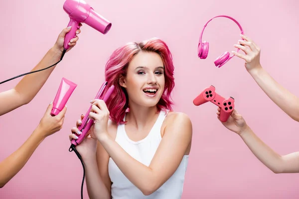 KYIV, UCRANIA - 29 de julio de 2020: mujer joven con el pelo colorido usando alisador cerca de los auriculares, crema cosmética, joystick y secador de pelo en las manos aisladas en rosa — Stock Photo