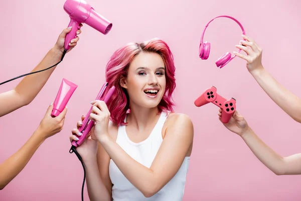 KYIV, UKRAINE - 29 JUILLET 2020 : jeune femme aux cheveux colorés utilisant redresseur près des écouteurs, crème cosmétique, joystick et sèche-cheveux dans les mains isolées sur rose — Photo de stock