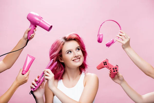 KYIV, UCRANIA - 29 de julio de 2020: mujer joven con el pelo colorido usando alisador cerca de los auriculares, crema cosmética, joystick y secador de pelo en las manos aisladas en rosa — Stock Photo