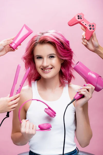 KYIV, UCRANIA - 29 de julio de 2020: mujer joven con el pelo colorido cerca de alisador, auriculares, crema cosmética, joystick y secador de pelo en las manos aisladas en rosa - foto de stock