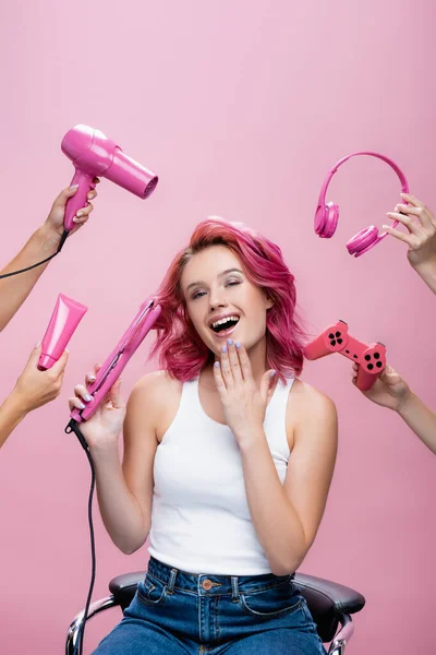 KYIV, UCRANIA - 29 de julio de 2020: mujer joven con el pelo colorido cerca de alisador, auriculares, crema cosmética, joystick y secador de pelo en las manos aisladas en rosa - foto de stock