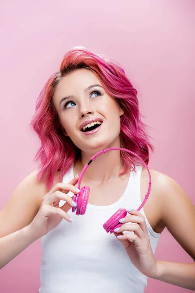 Mujer joven soñadora con el pelo colorido sosteniendo los auriculares y mirando hacia arriba aislado en rosa — Stock Photo