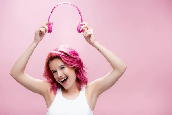 Збуджена молода жінка з барвистим волоссям, що тримає навушники над головою ізольовані на рожевому — Stock Photo