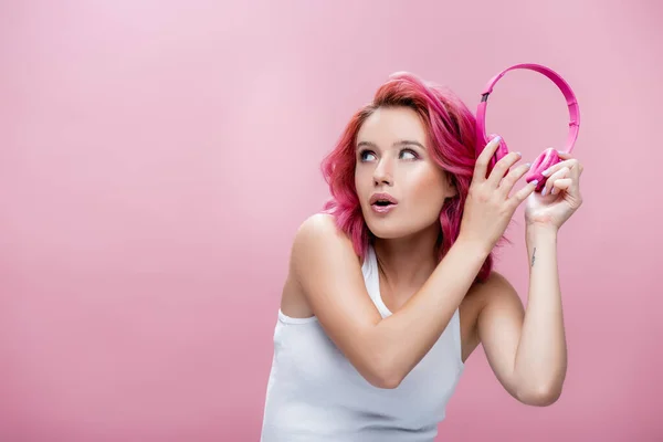 Mujer joven sorprendida con el pelo colorido celebración de auriculares cerca de la oreja aislado en rosa - foto de stock