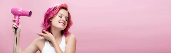 Mujer joven con el pelo colorido usando secador de pelo aislado en rosa, tiro panorámico - foto de stock
