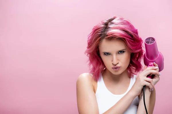 Jovem mulher séria com cabelo colorido segurando secador de cabelo isolado em rosa — Fotografia de Stock