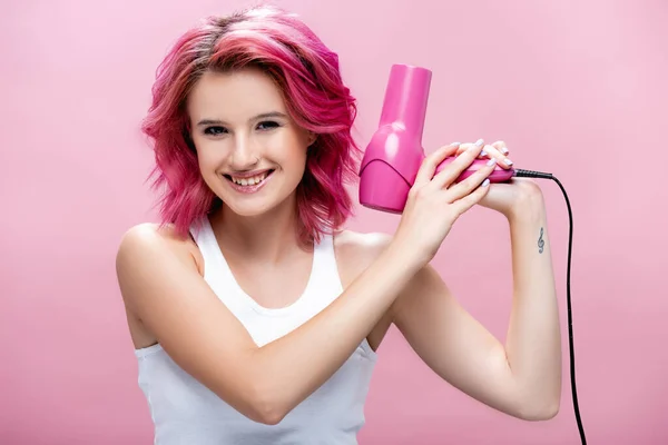 Junge Frau mit buntem Haar, Haartrockner in der Hand und isoliert lächelnd auf rosa — Stockfoto