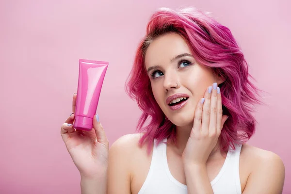Jovem sonhadora com tubo de cabelo colorido segurando de creme cosmético isolado em rosa — Fotografia de Stock