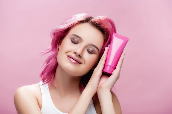 Jovem com cabelos coloridos e olhos fechados segurando tubo de creme cosmético isolado em rosa — Fotografia de Stock