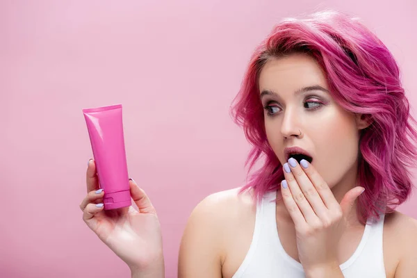 Scioccato giovane donna con i capelli colorati guardando tubo di crema cosmetica isolato su rosa — Foto stock