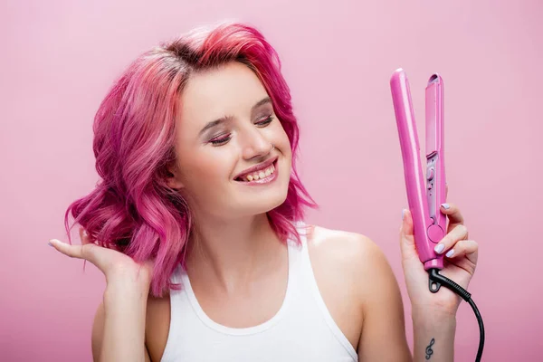 Junge Frau berührt bunte Haare und hält Glätteisen isoliert auf rosa — Stockfoto