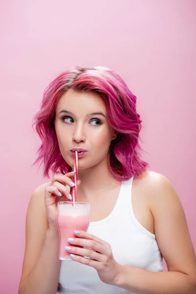 Jeune femme aux cheveux colorés buvant milkshake fraise de paille et regardant loin isolé sur rose — Photo de stock