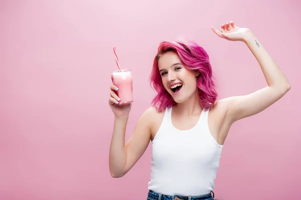 Young woman with colorful hair holding strawberry milkshake in glass with drinking straw and dancing isolated on pink — Stock Photo