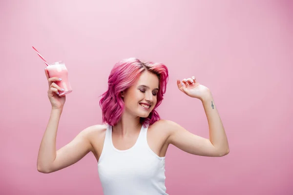 Young woman with colorful hair holding strawberry milkshake in glass with drinking straw and dancing isolated on pink — Stock Photo