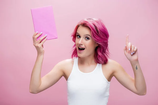 Aufgeregte junge Frau mit buntem Haar hält Buch in der Hand und zeigt Ideengeste isoliert auf rosa — Stockfoto