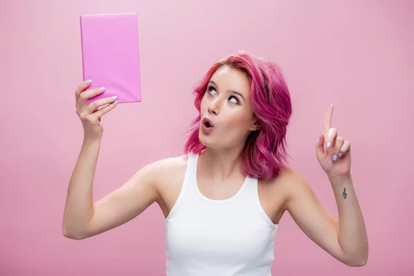 Surprised young woman with colorful hair holding book and showing idea gesture isolated on pink — Stock Photo