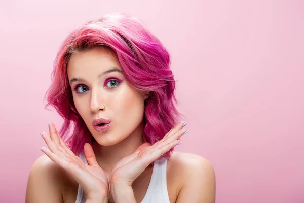 Mujer joven sorprendida con el pelo colorido y maquillaje posando con las manos cerca de la cara aislada en rosa - foto de stock