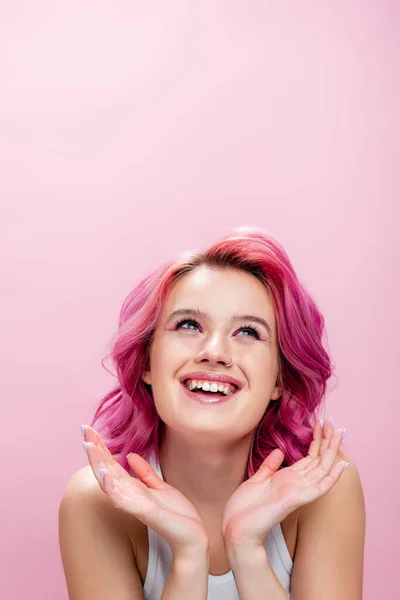 Mujer joven con el pelo colorido y maquillaje posando con las manos cerca de la cara y sonriendo aislado en rosa - foto de stock