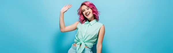 Молода жінка з рожевим волоссям махає рукою на синьому фоні, панорамний знімок — стокове фото