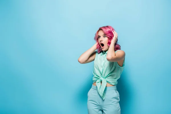 Schockierte junge Frau mit rosa Haaren und offenem Mund, die den Kopf auf blauem Hintergrund hält — Stockfoto