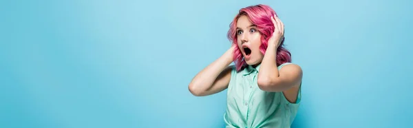 Jeune femme choquée aux cheveux roses et à la bouche ouverte tenant la tête sur fond bleu, prise de vue panoramique — Photo de stock