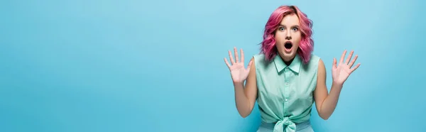 Jeune femme choquée aux cheveux roses et à la bouche ouverte montrant les mains sur fond bleu, prise de vue panoramique — Photo de stock