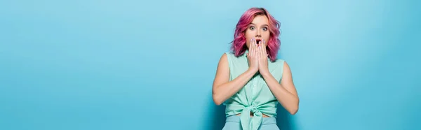 Шокированная молодая женщина с розовыми волосами, покрывающими рот на синем фоне, панорамный снимок — стоковое фото