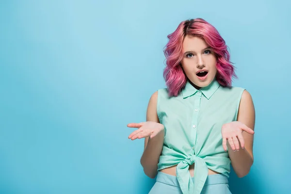 Здивована молода жінка з рожевим волоссям і відкритим ротом, що показує жест згинання на синьому фоні — стокове фото