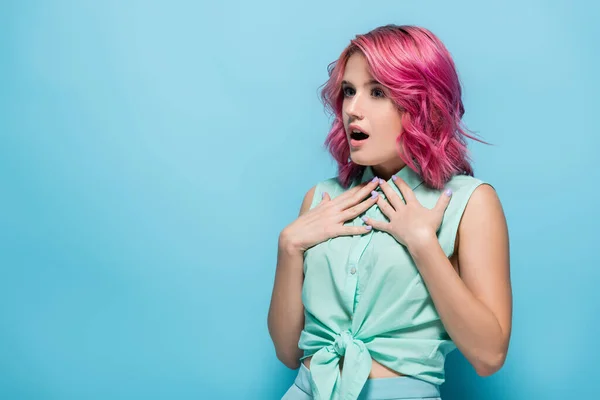 Здивована молода жінка з рожевим волоссям і відкритим ротом торкається обличчя на синьому фоні — стокове фото