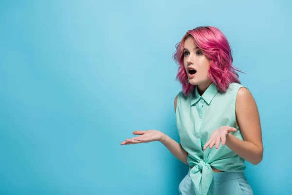Surpresa jovem mulher com cabelo rosa e boca aberta mostrando gesto de encolher de ombros no fundo azul — Fotografia de Stock