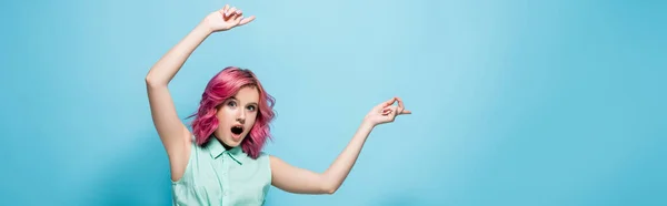 Chocado jovem mulher com cabelo rosa apontando para o lado no fundo azul, tiro panorâmico — Fotografia de Stock