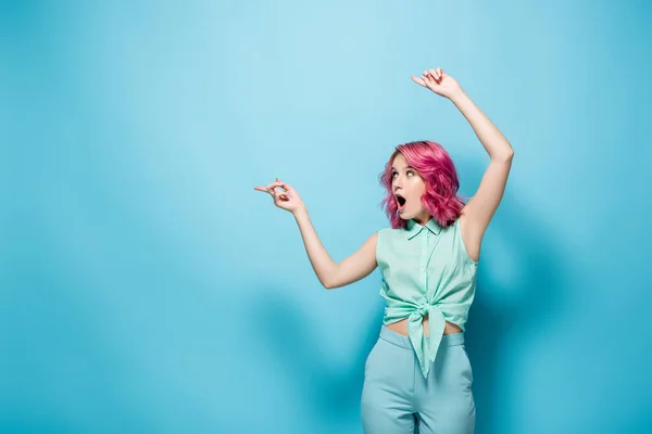 Choqué jeune femme avec les cheveux roses pointant de côté sur fond bleu — Photo de stock