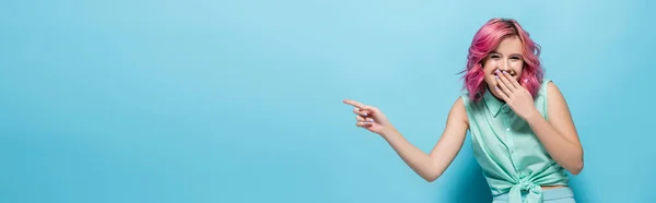 Jeune femme aux cheveux roses pointant du doigt et riant sur fond bleu, plan panoramique — Photo de stock