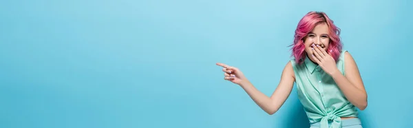 Jovem mulher com cabelo rosa apontando para o lado e rindo no fundo azul, tiro panorâmico — Fotografia de Stock