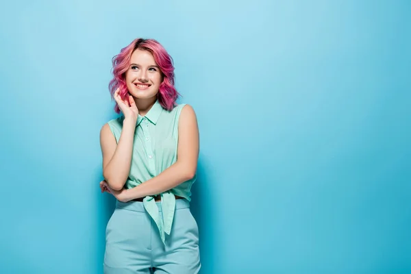 Mujer joven soñadora con el pelo rosa sonriendo y mirando hacia otro lado sobre fondo azul - foto de stock