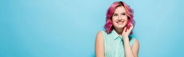 Jeune femme rêveuse aux cheveux roses souriant et regardant loin sur fond bleu, prise de vue panoramique — Photo de stock