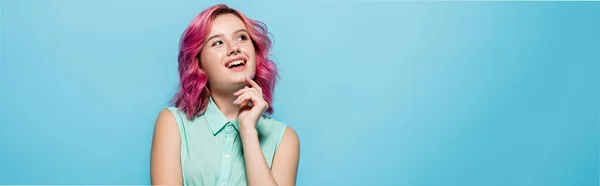 Mujer joven soñadora con el pelo rosa sonriendo y mirando hacia otro lado aislado en azul, plano panorámico - foto de stock