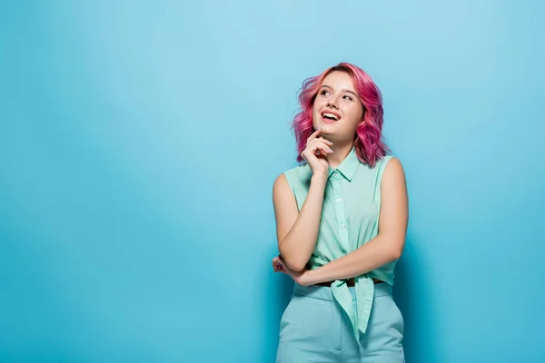 Verträumte junge Frau mit rosa Haaren, die vor blauem Hintergrund lächelt und wegschaut — Stockfoto