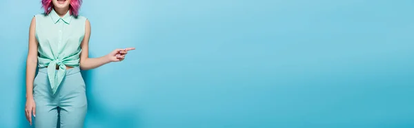 Cropped view of young woman with pink hair pointing aside on blue background, panoramic shot — Stock Photo