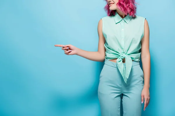 Vue recadrée de jeune femme aux cheveux roses pointant de côté sur fond bleu — Photo de stock