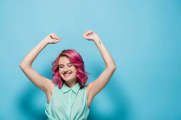 Young woman with pink hair and hands in air smiling on blue background — Stock Photo
