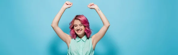 Young woman with pink hair and hands in air smiling on blue background, panoramic shot — Stock Photo