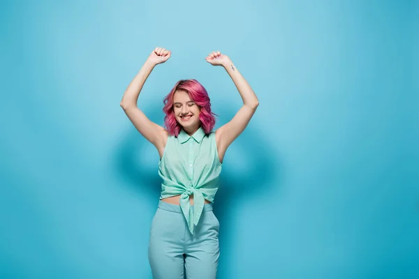 Young woman with pink hair and hands in air smiling on blue background — Stock Photo