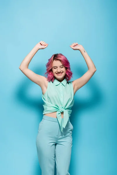 Junge Frau mit rosa Haaren und Händen in der Luft, die auf blauem Hintergrund lächeln — Stockfoto