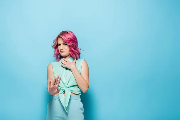 Mujer joven confundida con el pelo rosa mostrando rechazo sobre fondo azul - foto de stock