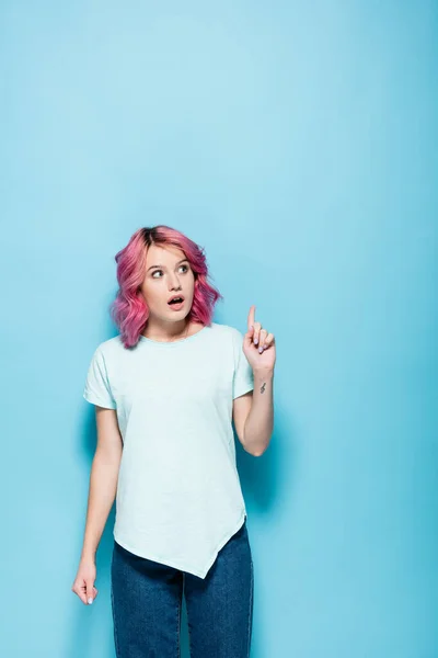 Mujer joven sorprendida con el pelo rosa apuntando hacia arriba en el fondo azul - foto de stock