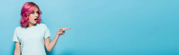 Surprise jeune femme aux cheveux roses pointant de côté sur fond bleu, plan panoramique — Photo de stock