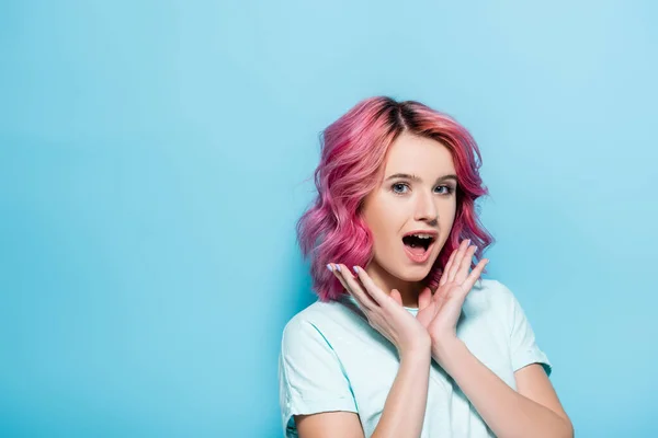 Mujer joven sorprendida con el pelo rosa y las manos cerca de la cara sobre fondo azul - foto de stock