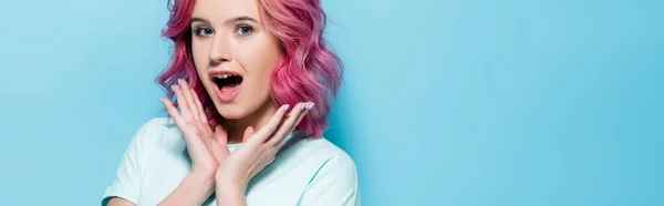 Удивлена молодая женщина с розовыми волосами и руками возле лица на синем фоне, панорамный снимок — стоковое фото